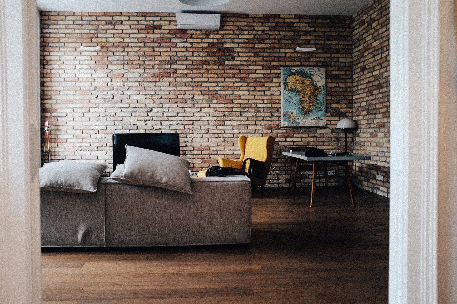 Living room with exposed brick wall, gray sofa, yellow chair, and small dining table by a window