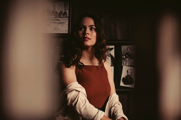 Woman in a red top and white jacket, standing in a dim room with framed pictures on the wall.