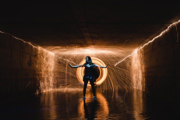 Person standing in a glowing tunnel, arms outstretched, with warm orange light and wet reflections.