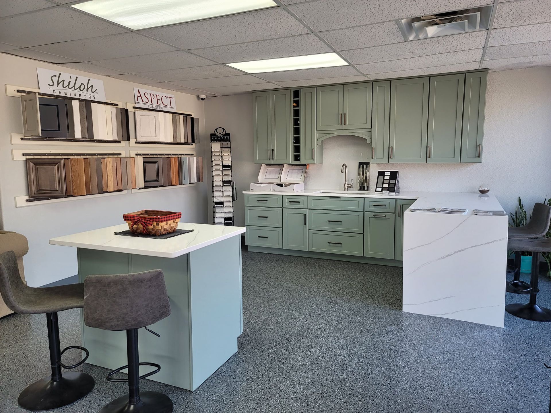 Kitchen showroom featuring cabinets, countertops, and sample materials, with island seating and speckled flooring.