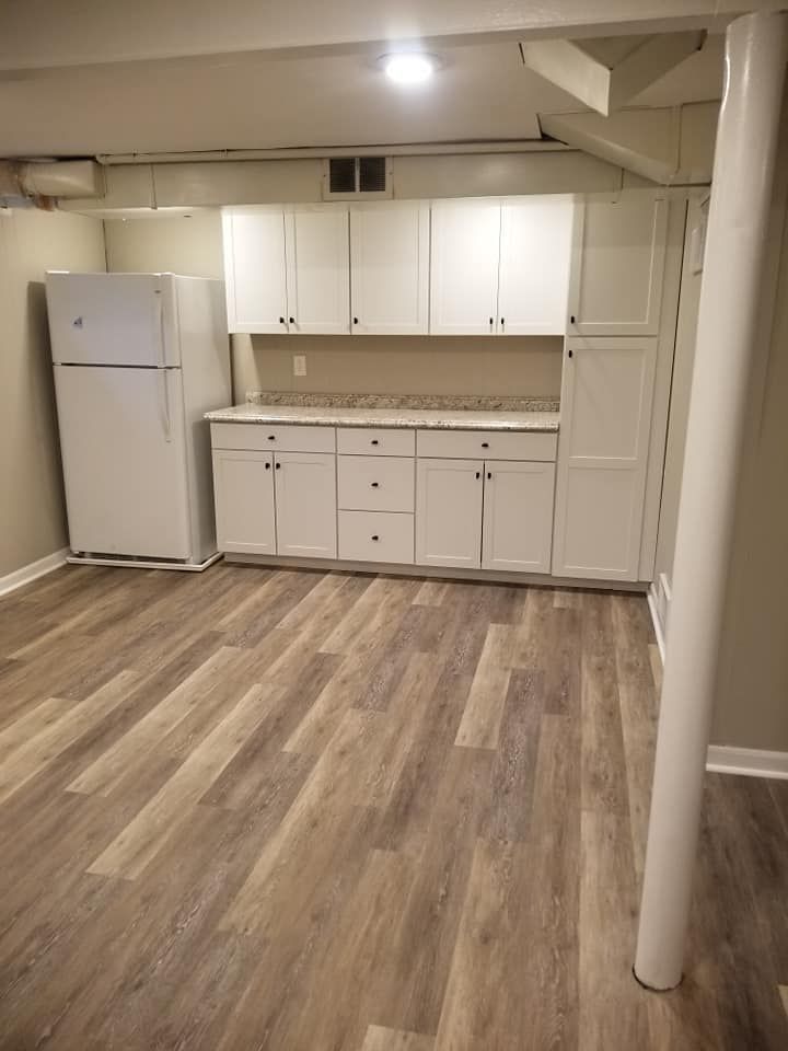 Basement kitchen with white cabinets, countertop, refrigerator, and wood-look flooring.