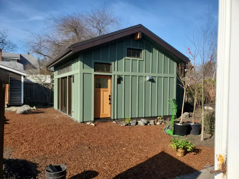 A green shed with a brown roof is in the backyard of a house.