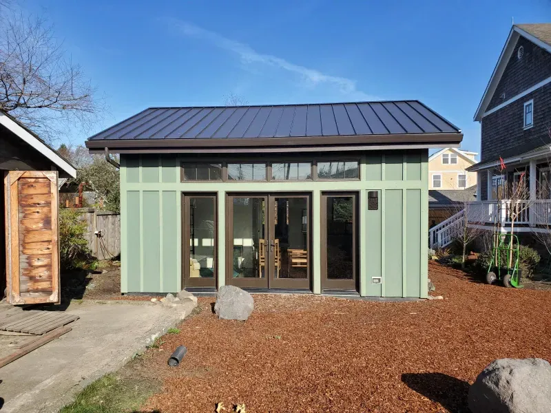 A small green house with a black roof is sitting in the middle of a yard next to a house.