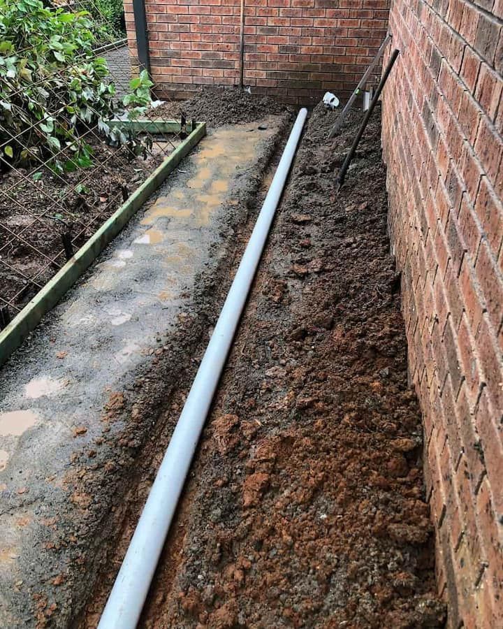 A White Pipe Is Laying In The Dirt Next To A Brick Wall — ARK Plumbing & Gasfitting In Berrima, NSW