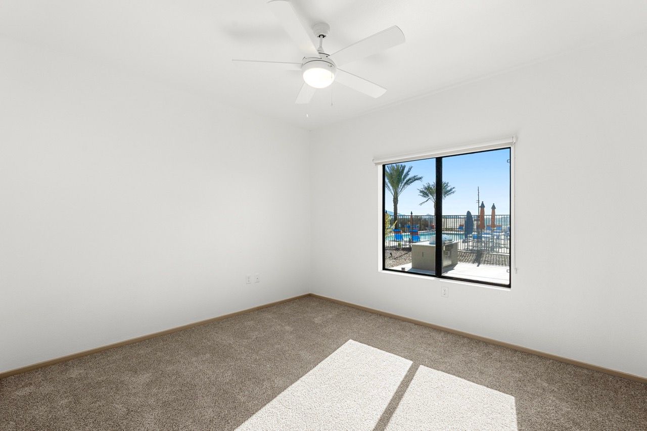 Vacant bedroom with beige carpet, white walls, a ceiling fan, and a window overlooking the pool.