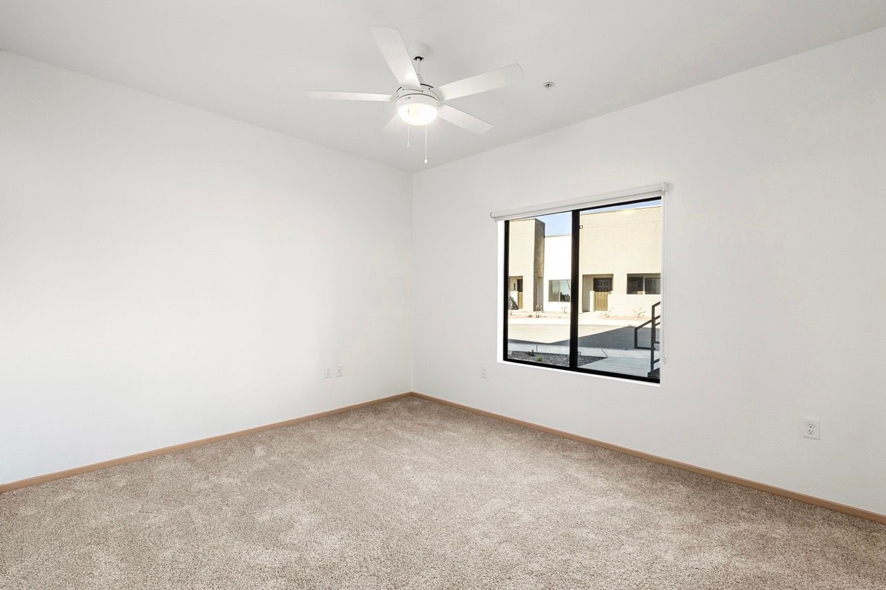 Empty bedroom with beige carpet, white walls, window, and ceiling fan.