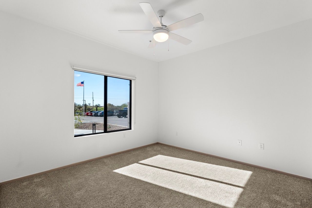 Empty bedroom with white walls, beige carpet, a window, and a ceiling fan.