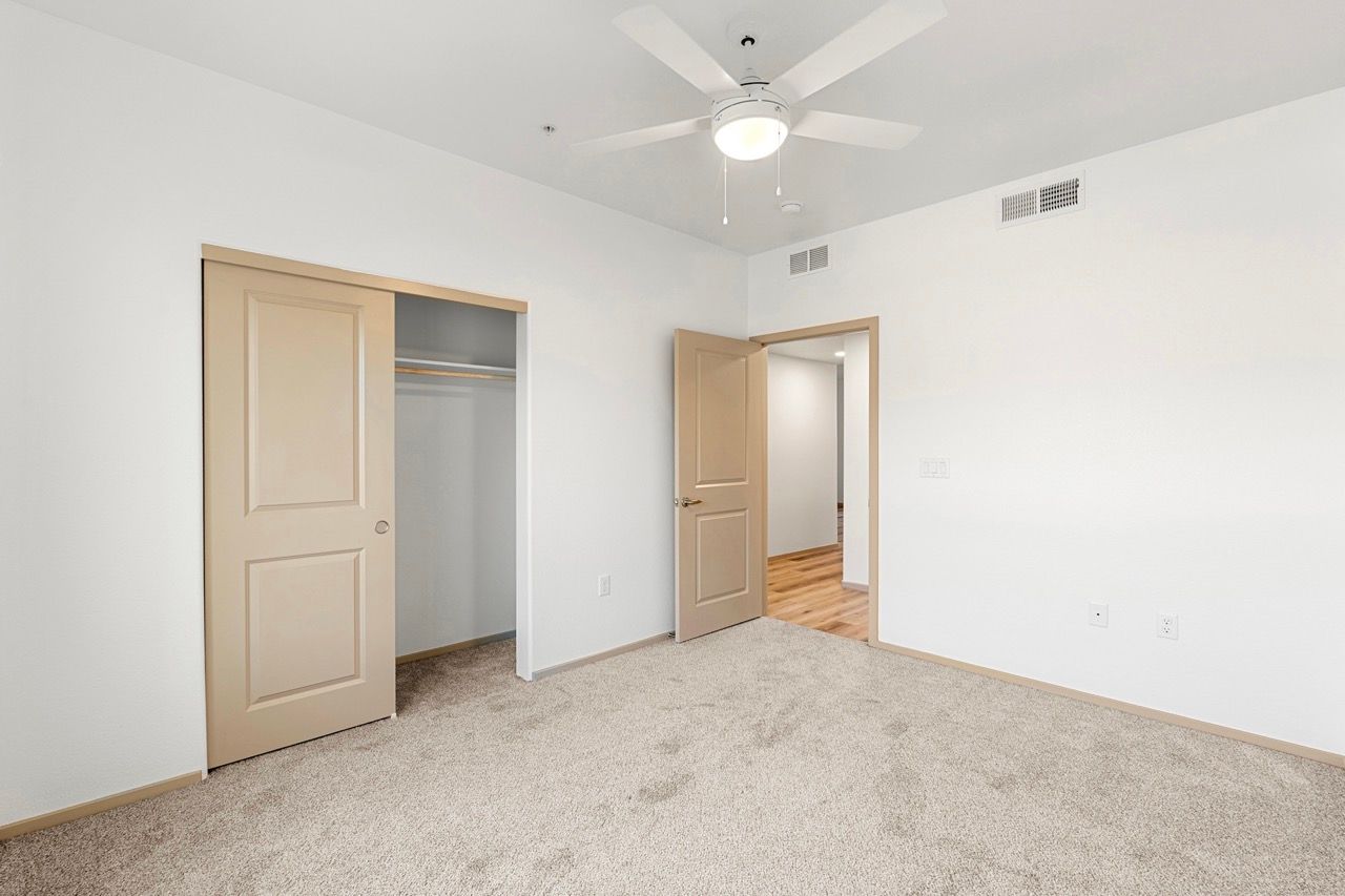 Empty bedroom with beige closet doors, ceiling fan, and carpet.