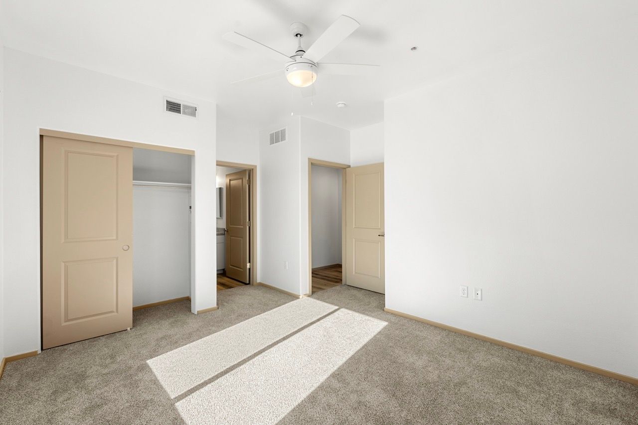 Bright bedroom with neutral walls, carpet, a ceiling fan, and beige closet doors.