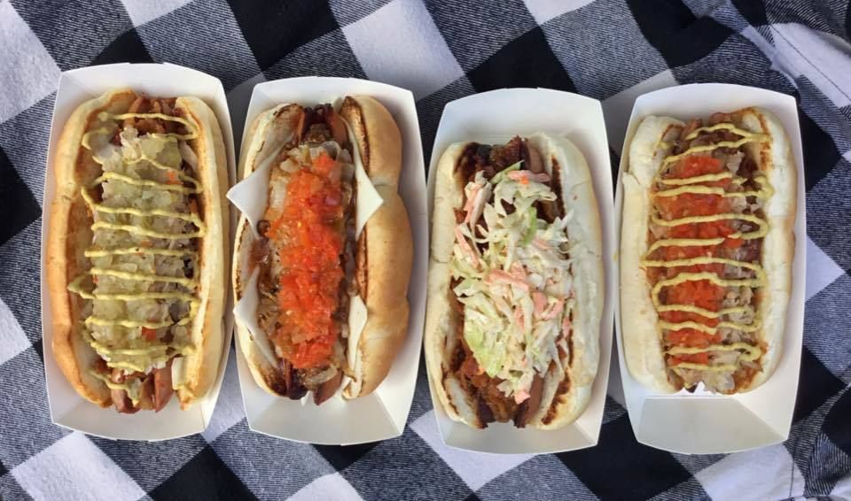 Four hot dogs with different toppings are sitting on a checkered blanket.