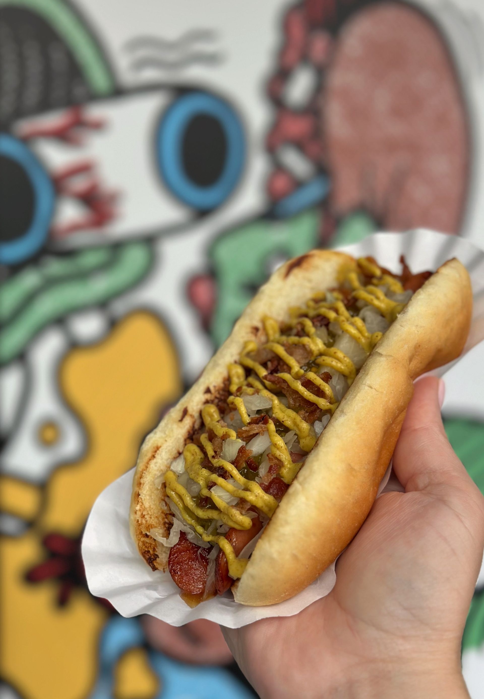 A person is holding a hot dog with mustard and onions on a paper plate.