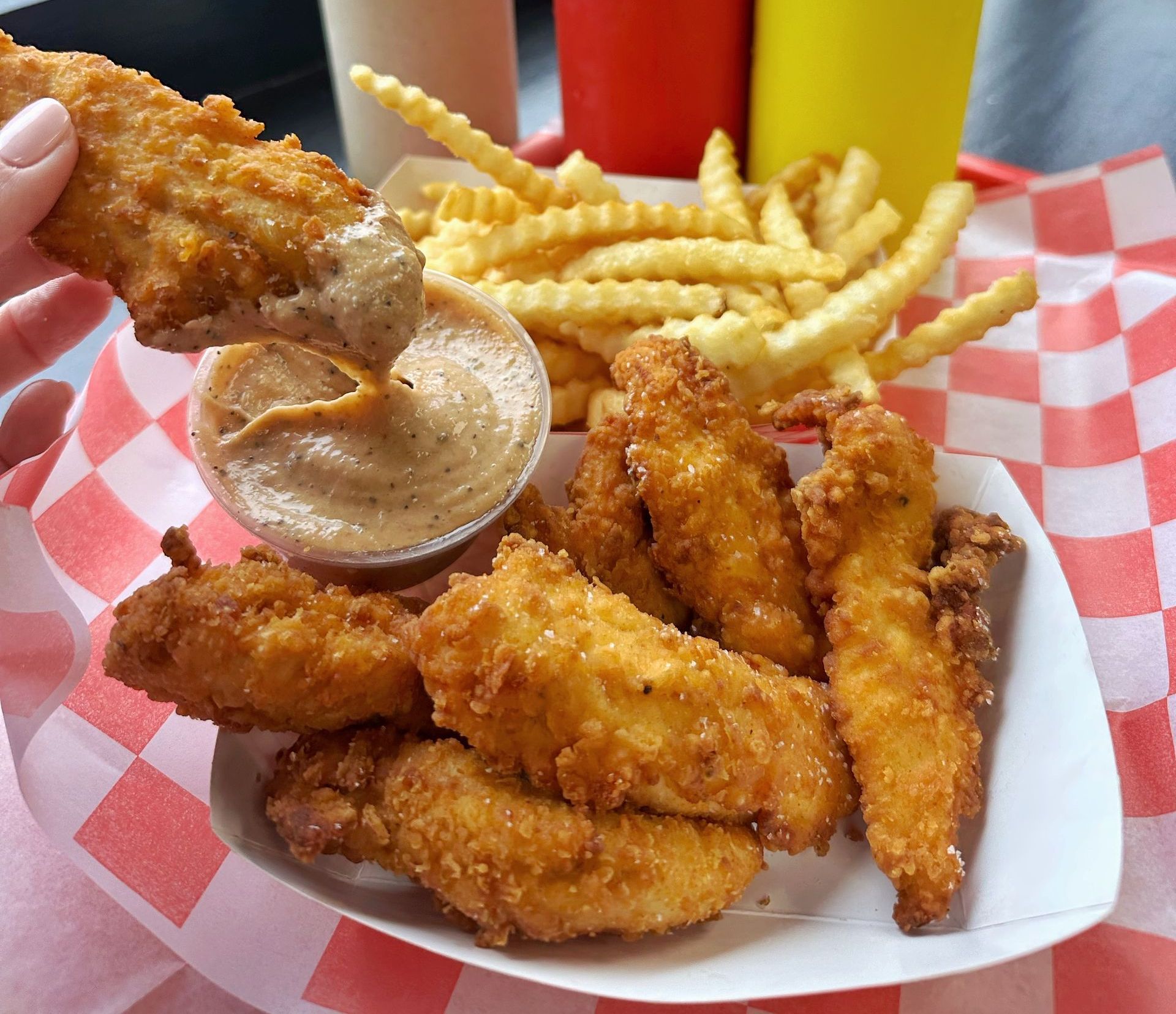 A person is dipping a piece of chicken into a sauce next to french fries.