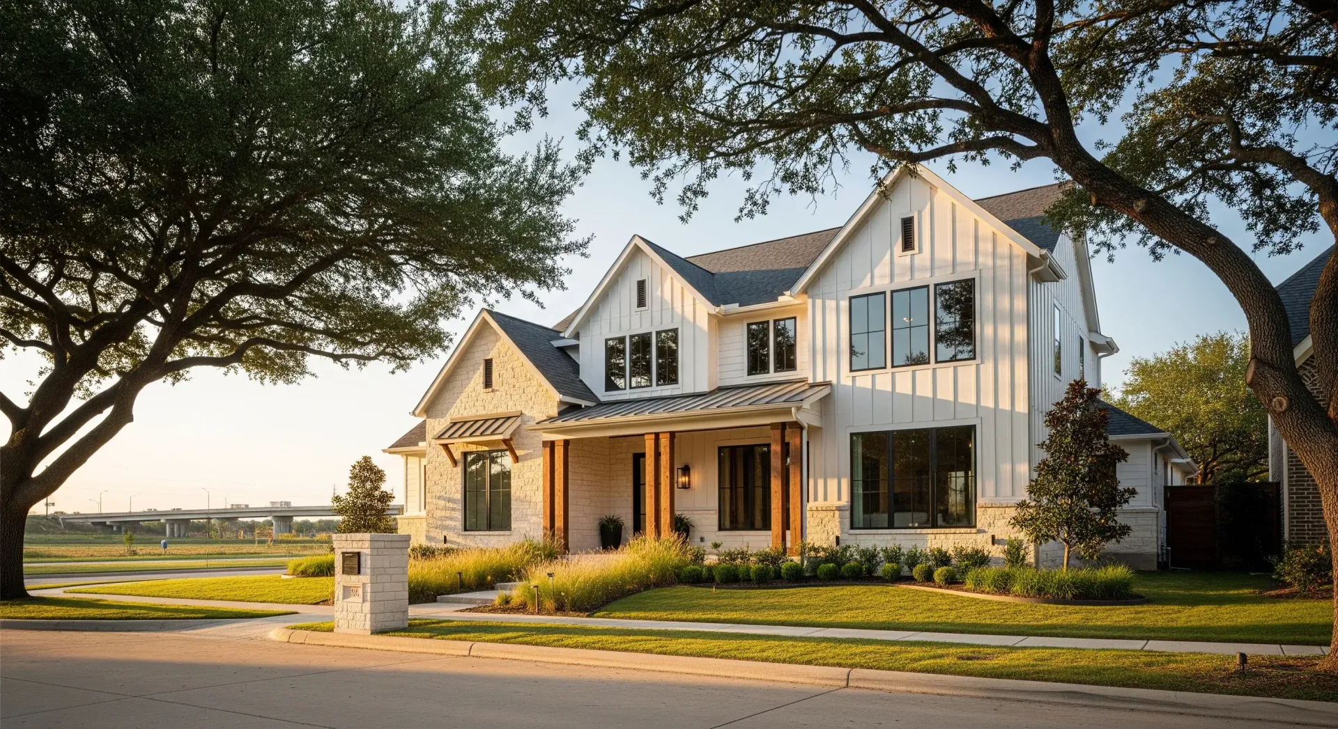 A two-story farmhouse-style home with white vertical siding and stone accents, framed by large trees at golden hour.