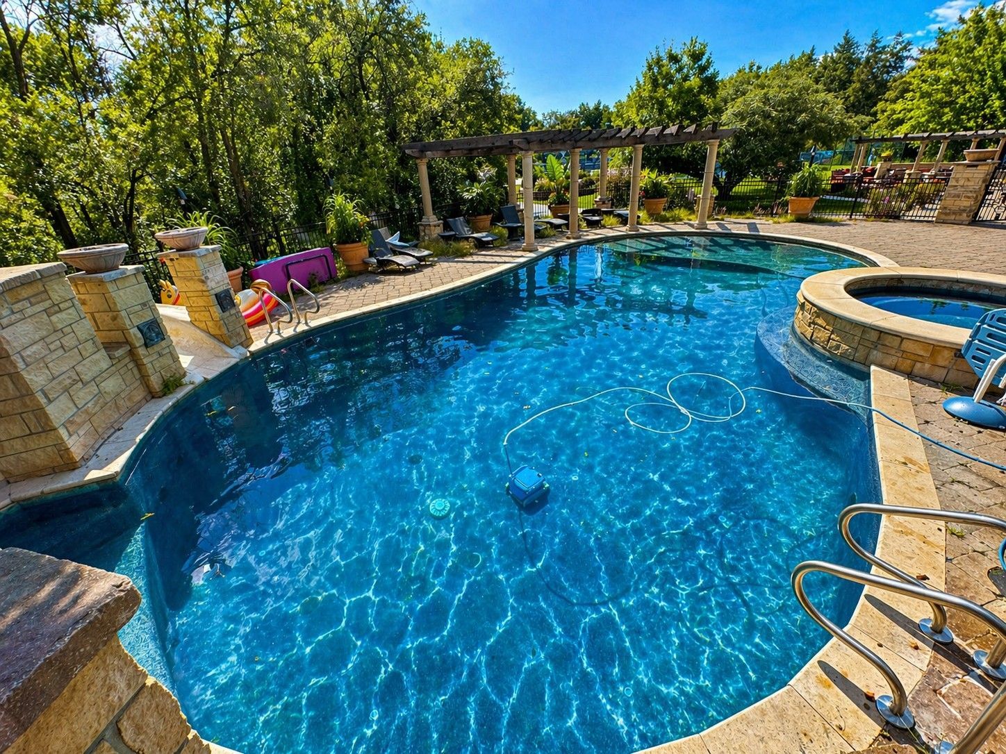Backyard pool with blue water, stone deck, spa, and trees under a sunny sky
