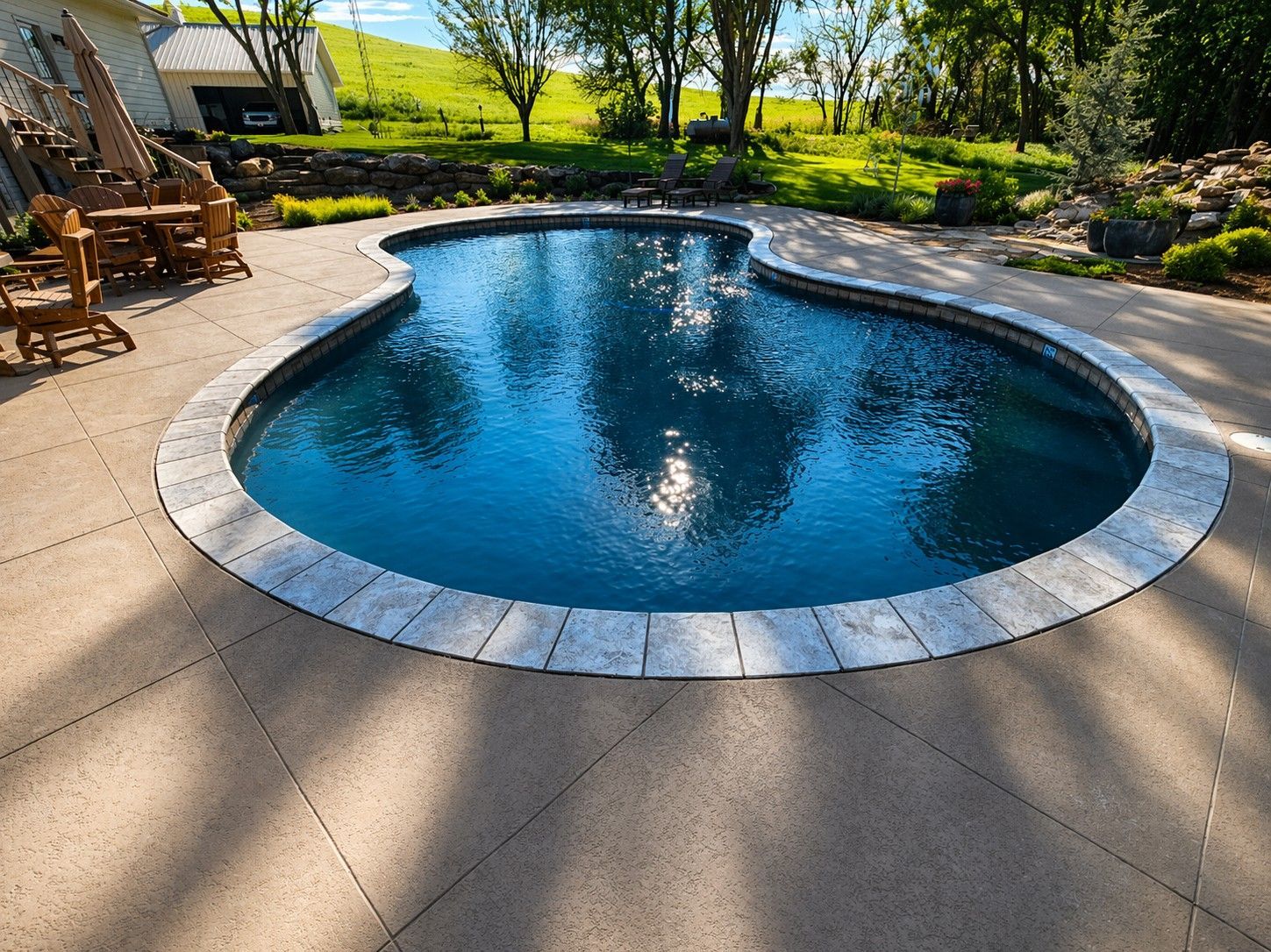 Curved backyard swimming pool with blue water, surrounded by stone patio and green trees in bright sunlight
