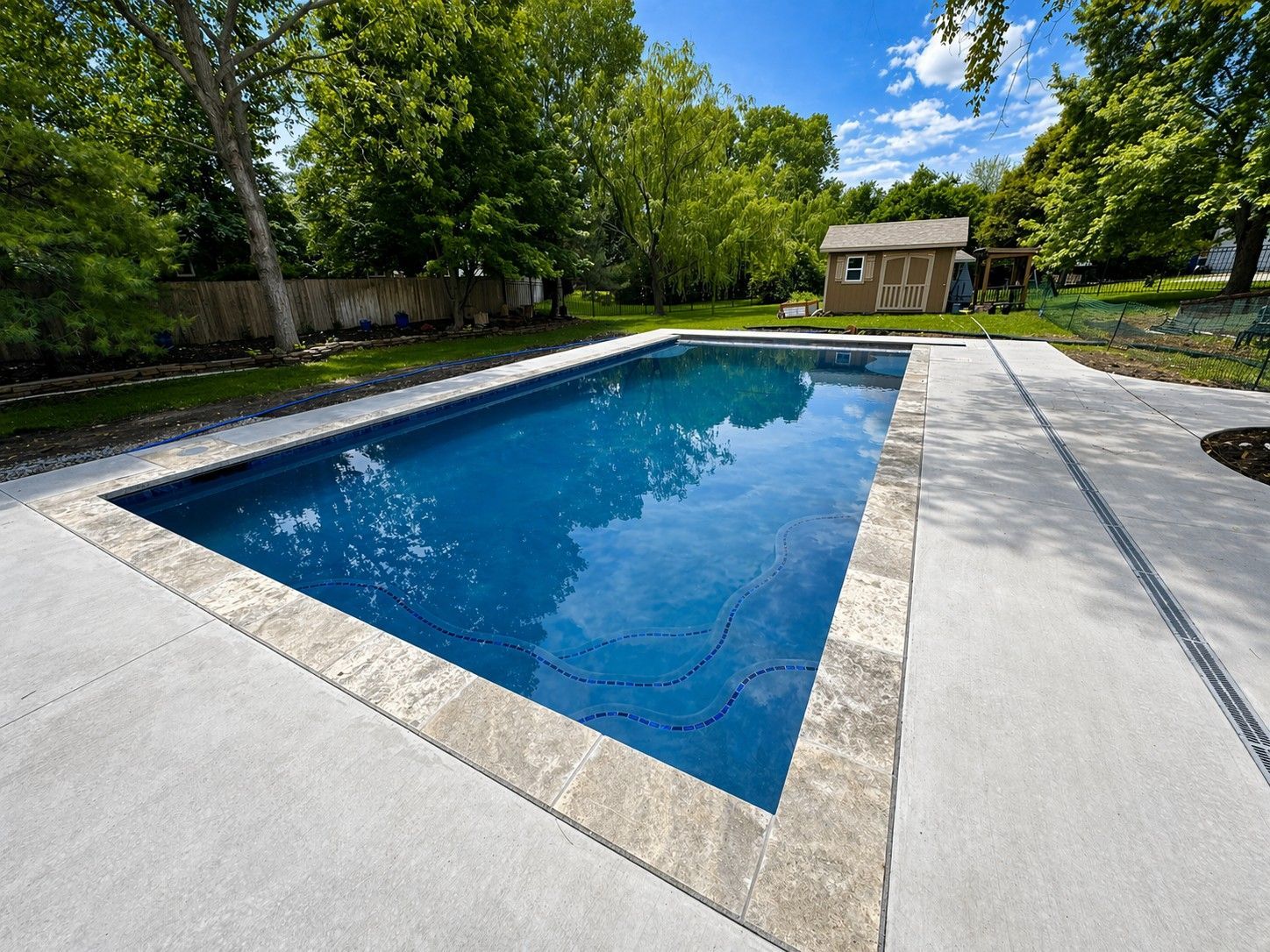 Backyard swimming pool with blue water, concrete deck, and trees in the background