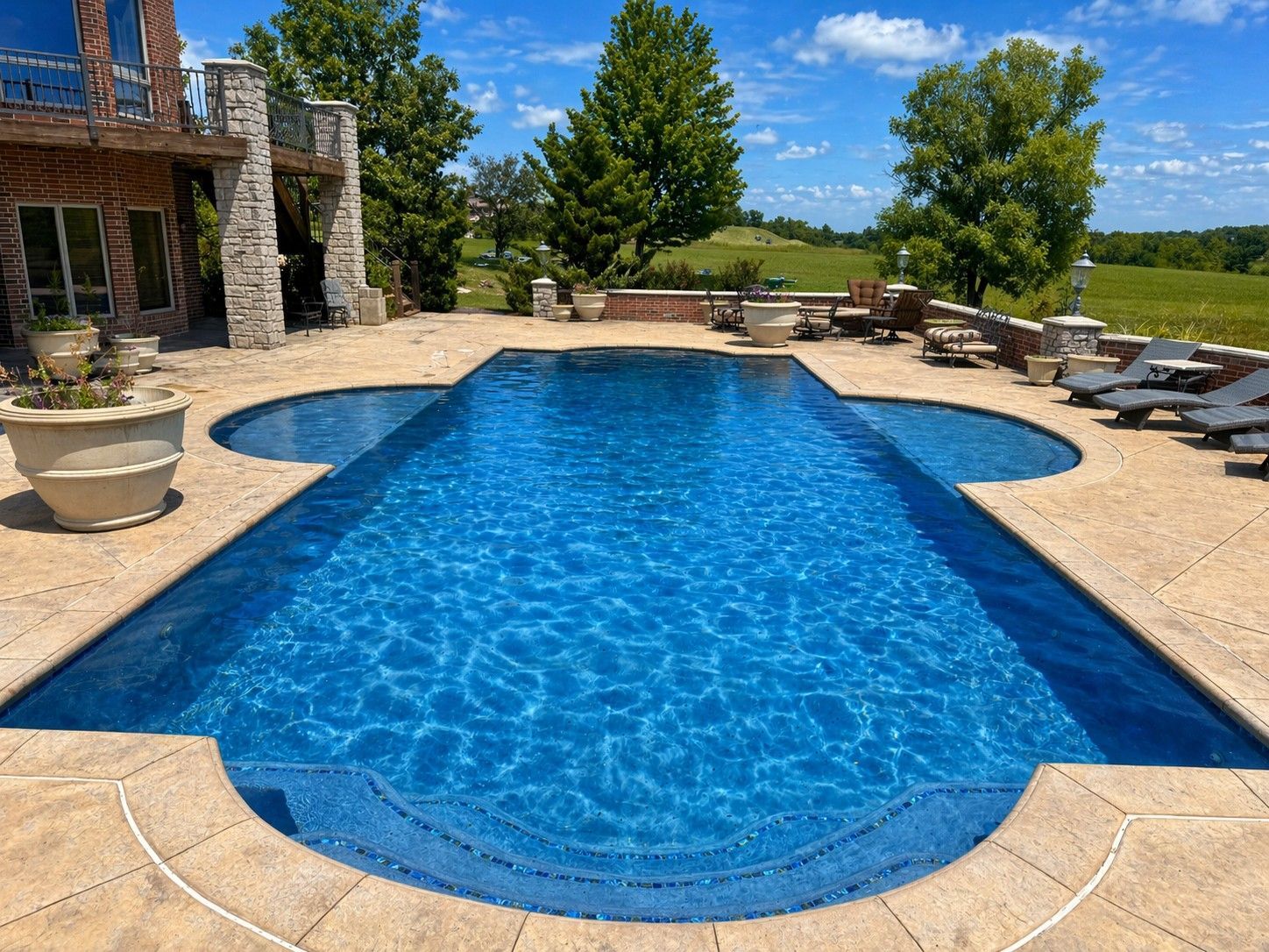 Blue backyard pool with stone patio, lounge chairs, and a stone house under a clear sky