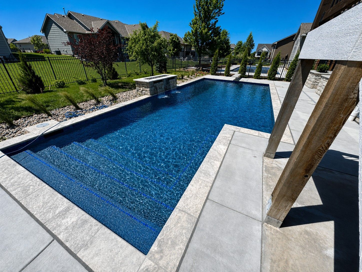 Backyard rectangular swimming pool with blue water, stone deck, and landscaped yard under a clear sky