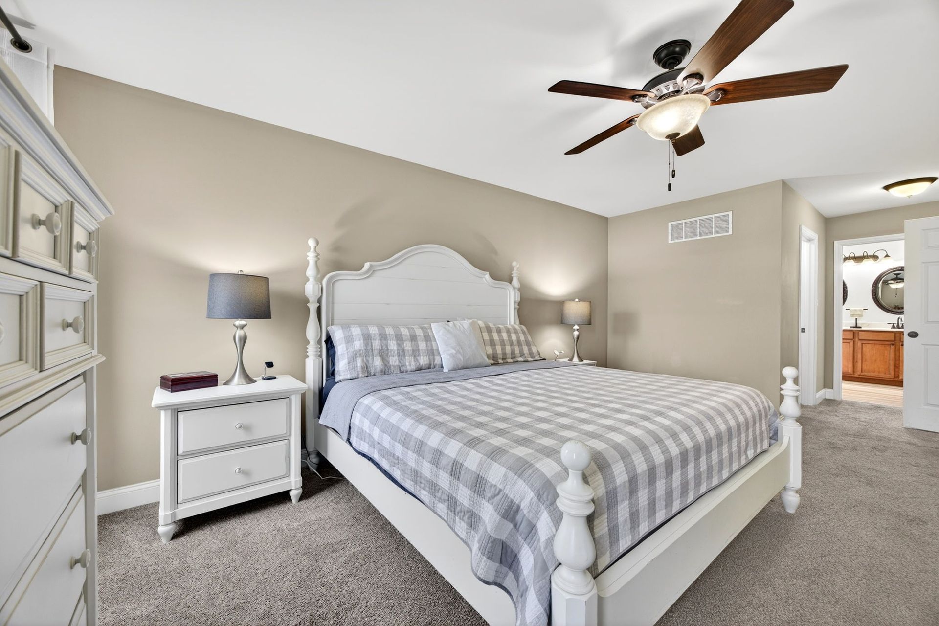 A beige bedroom featuring a white bed with a plaid comforter, a white nightstand, and a ceiling fan.