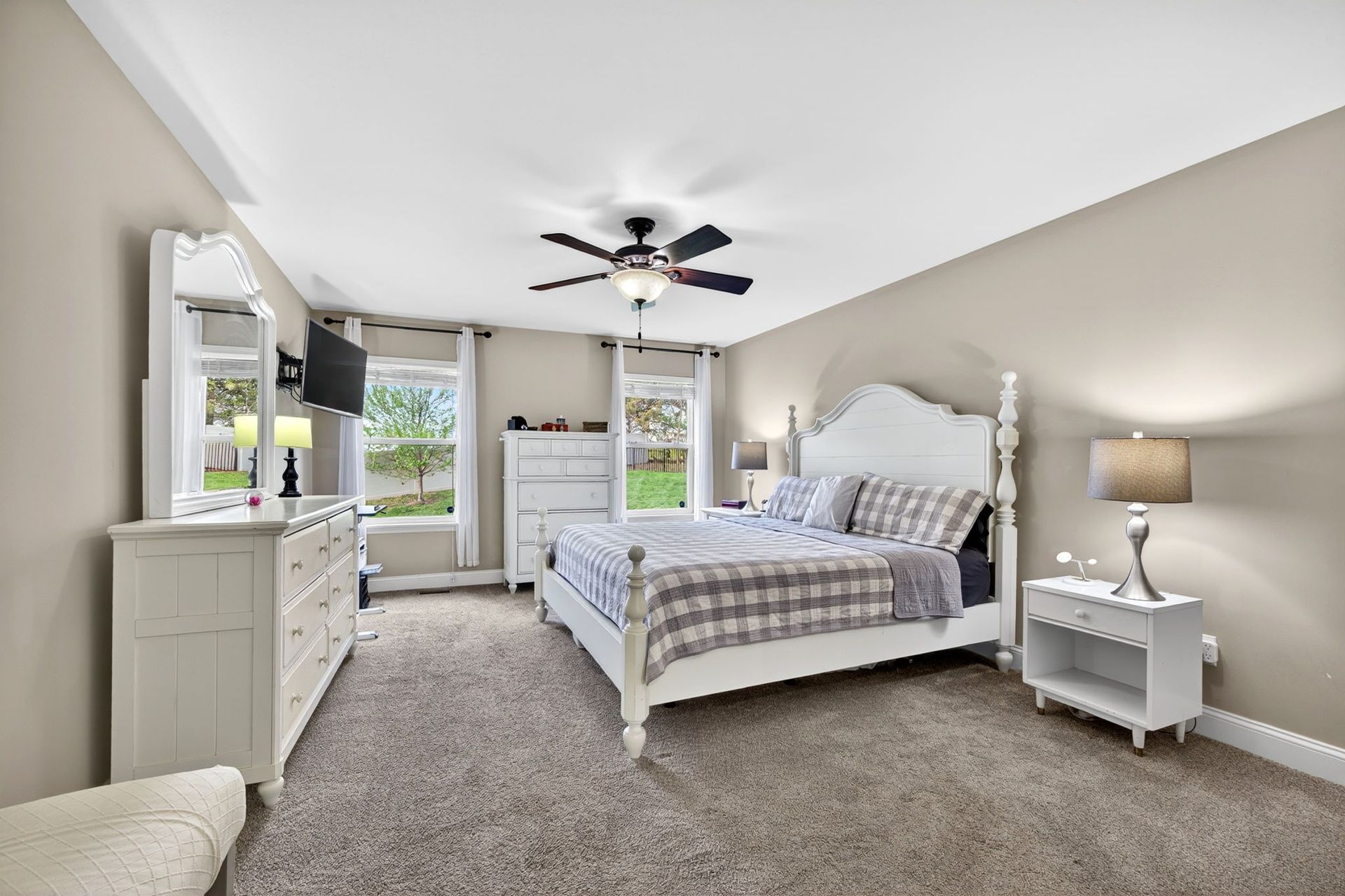 A bright bedroom with a white bed, white dresser with mirror, nightstand, and ceiling fan over gray wall-to-wall carpet.
