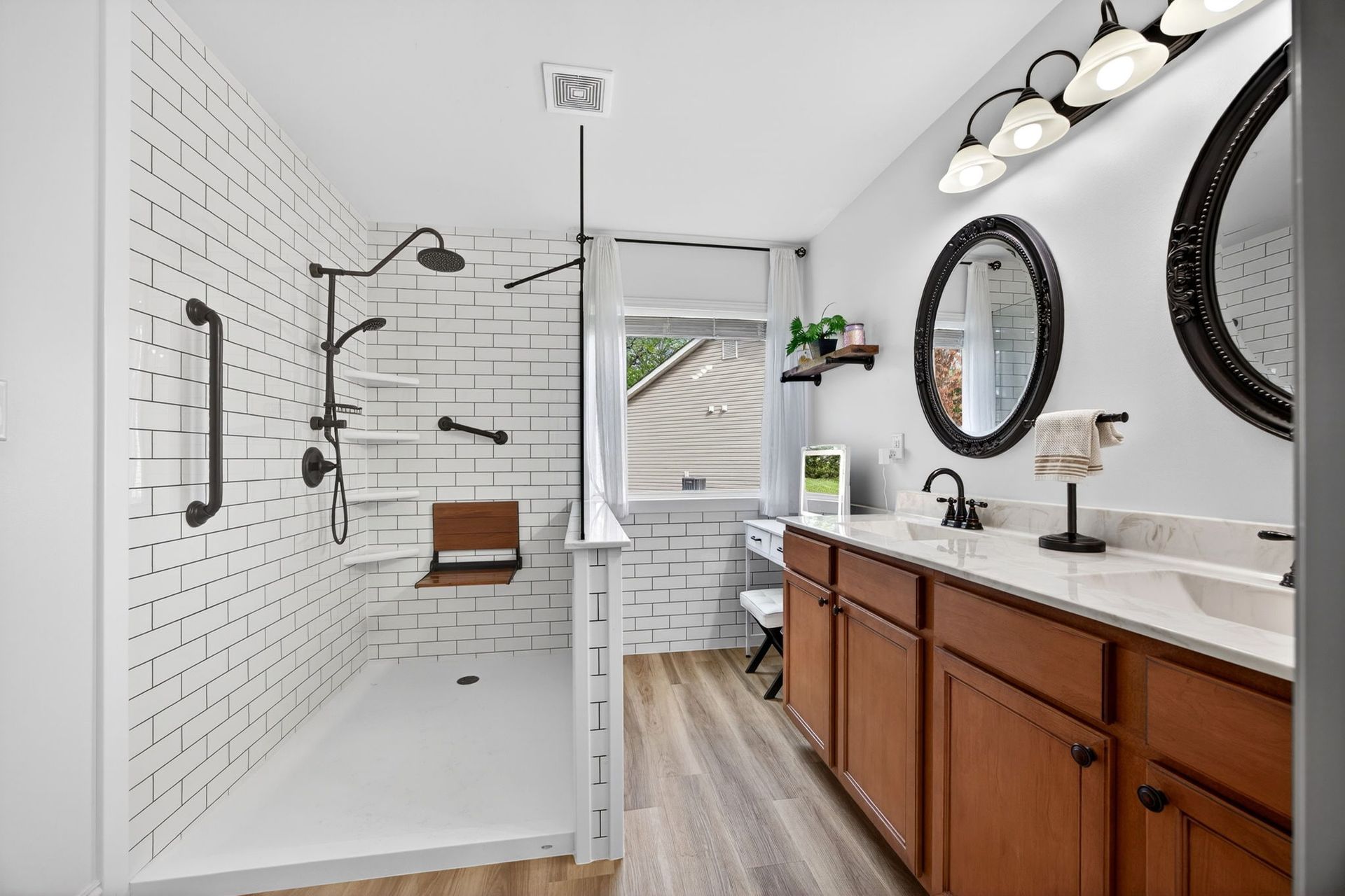 Accessible bathroom with a walk-in shower featuring a wooden seat, grab bars, white brick walls, and dual wood vanities.