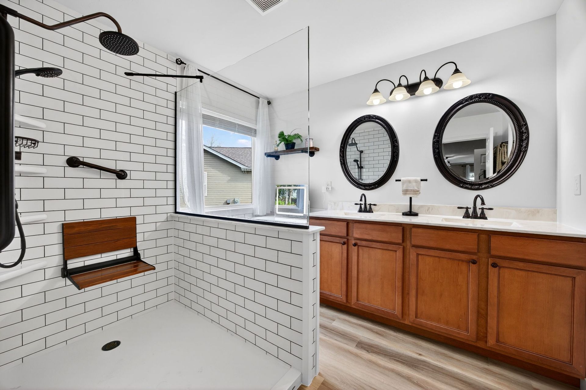 A modern bathroom with a white brick walk-in shower, wooden bench, double vanity with round mirrors, and wood-look floors.