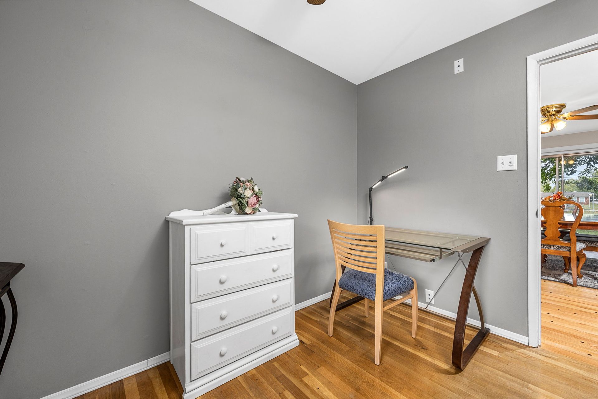 A room with a desk , chair , dresser and ceiling fan.