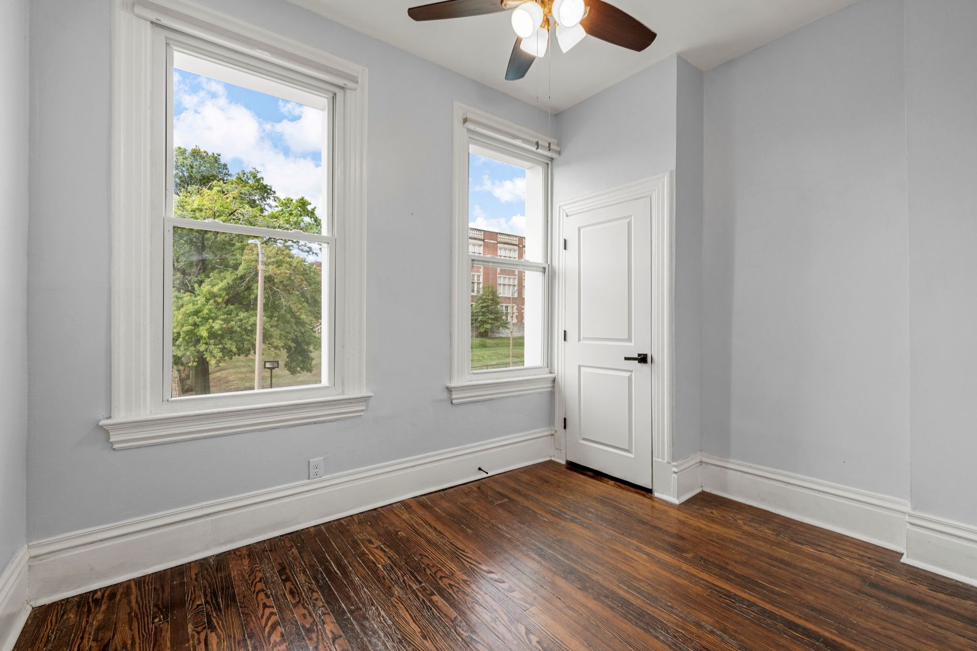Empty room with hardwood floors, two windows, and a white door.