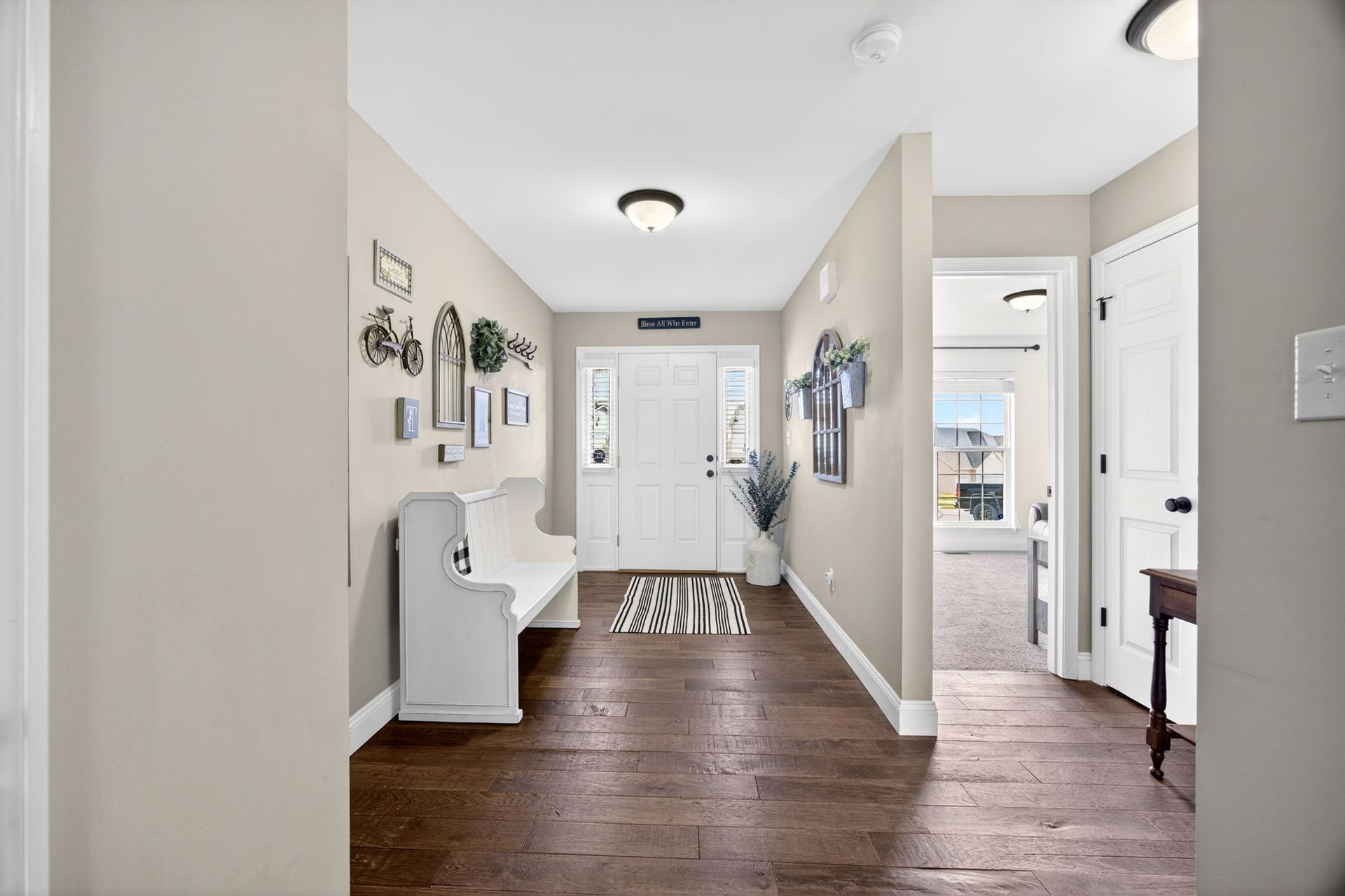 A hallway with hardwood floors leading to a white front door, a white bench, wall decor, and a view into another room.