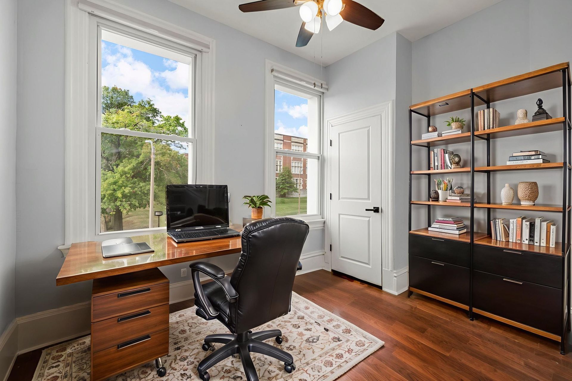 Home office with desk, chair, bookshelf, and windows overlooking trees.