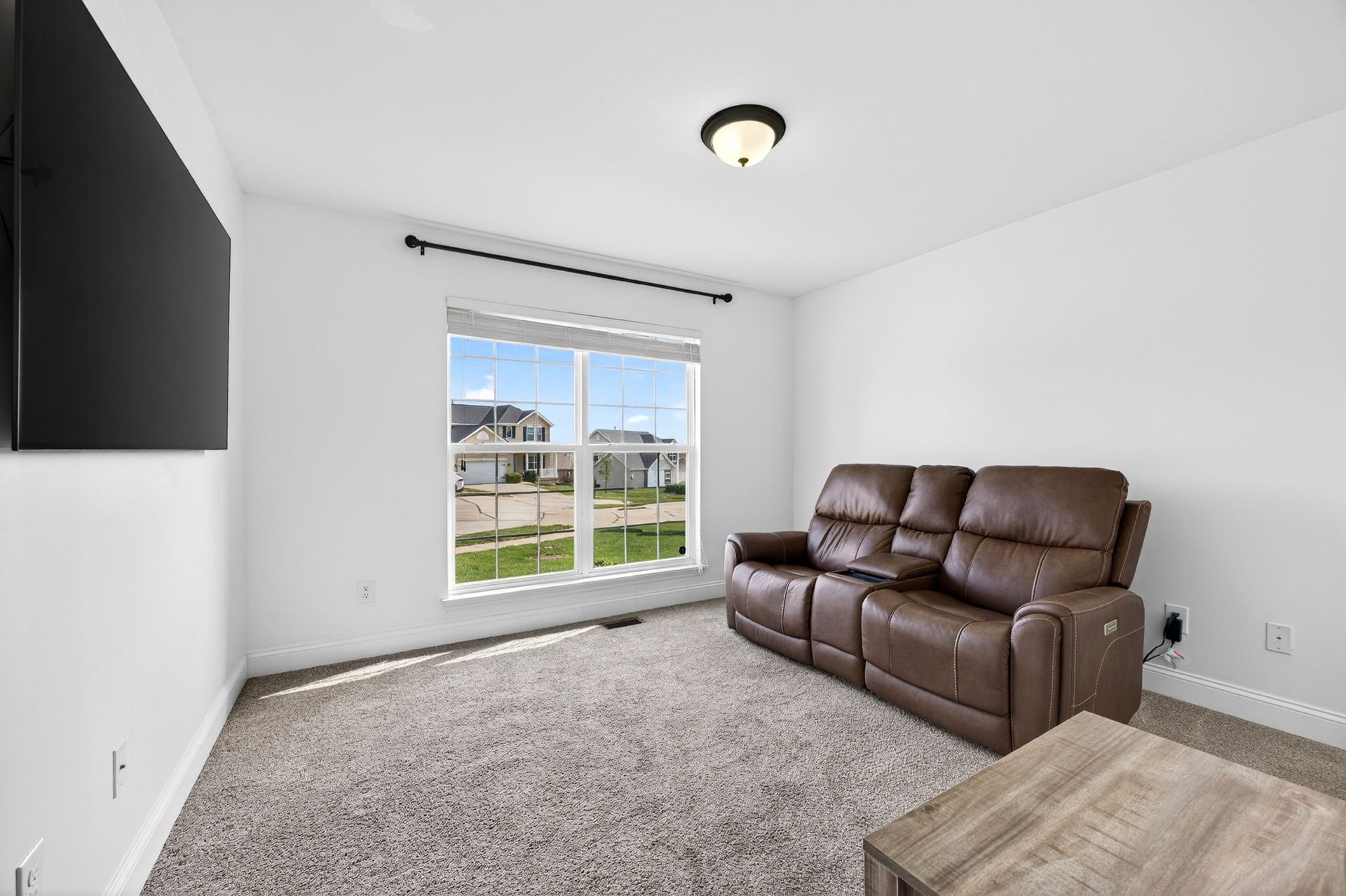 A sparsely furnished room with white walls, carpet, a brown leather reclining sofa, and a large wall-mounted television.