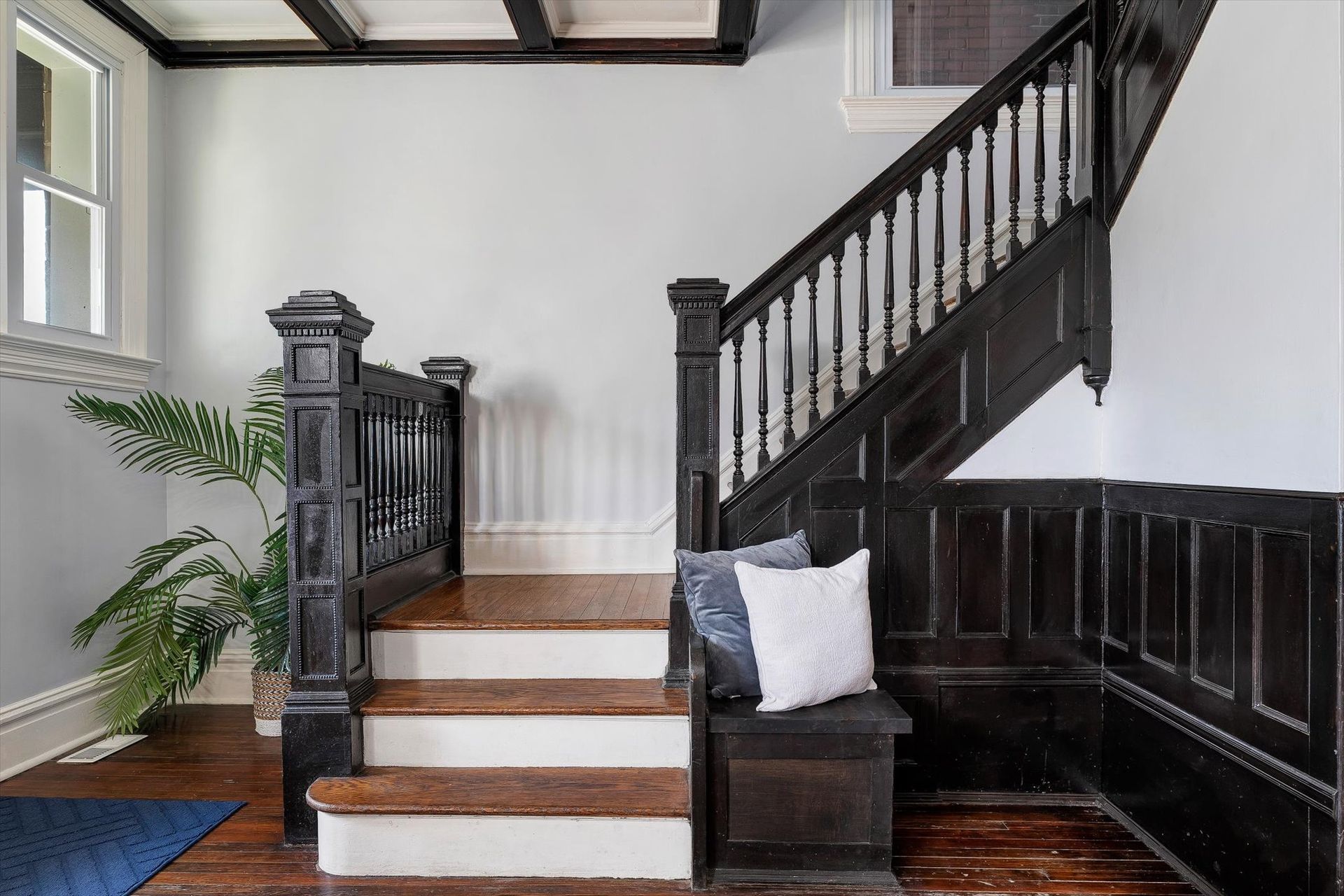 A wooden staircase with black trim, white walls, and a bench with pillows. A plant sits nearby.