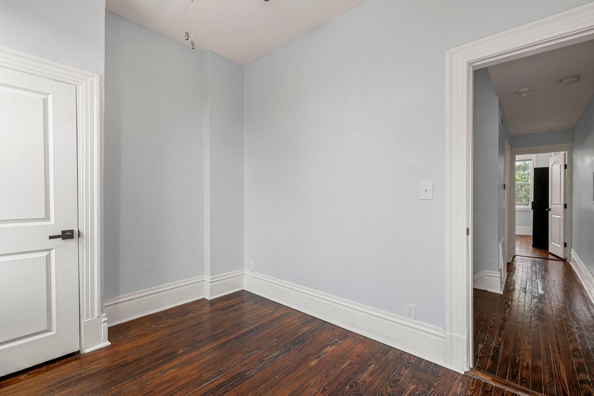 Empty room with light blue walls, white trim, and dark wood floors; doorway on the right.