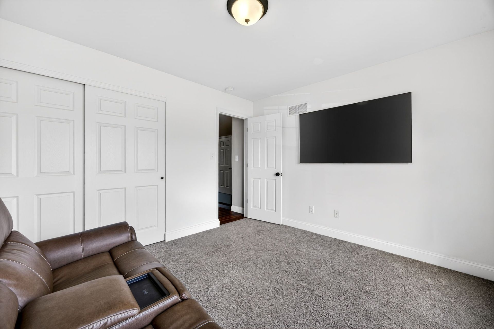 A cozy room features a dark brown leather recliner, a large wall-mounted TV, grey carpet, and white interior doors.