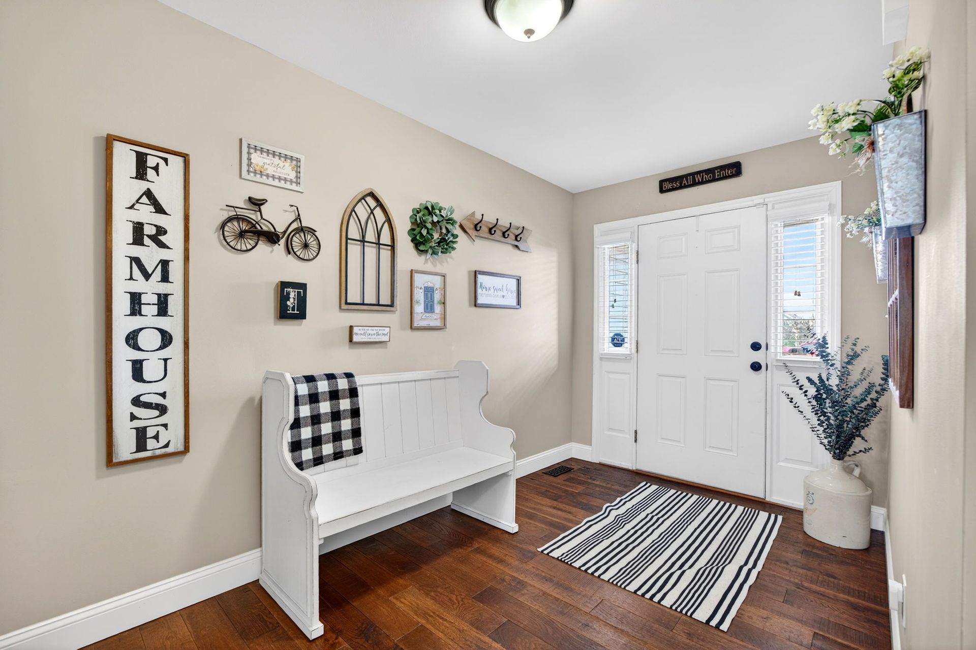 A farmhouse-style entryway with a white bench, wall decor, dark wood floors, and a white front door with sidelights.