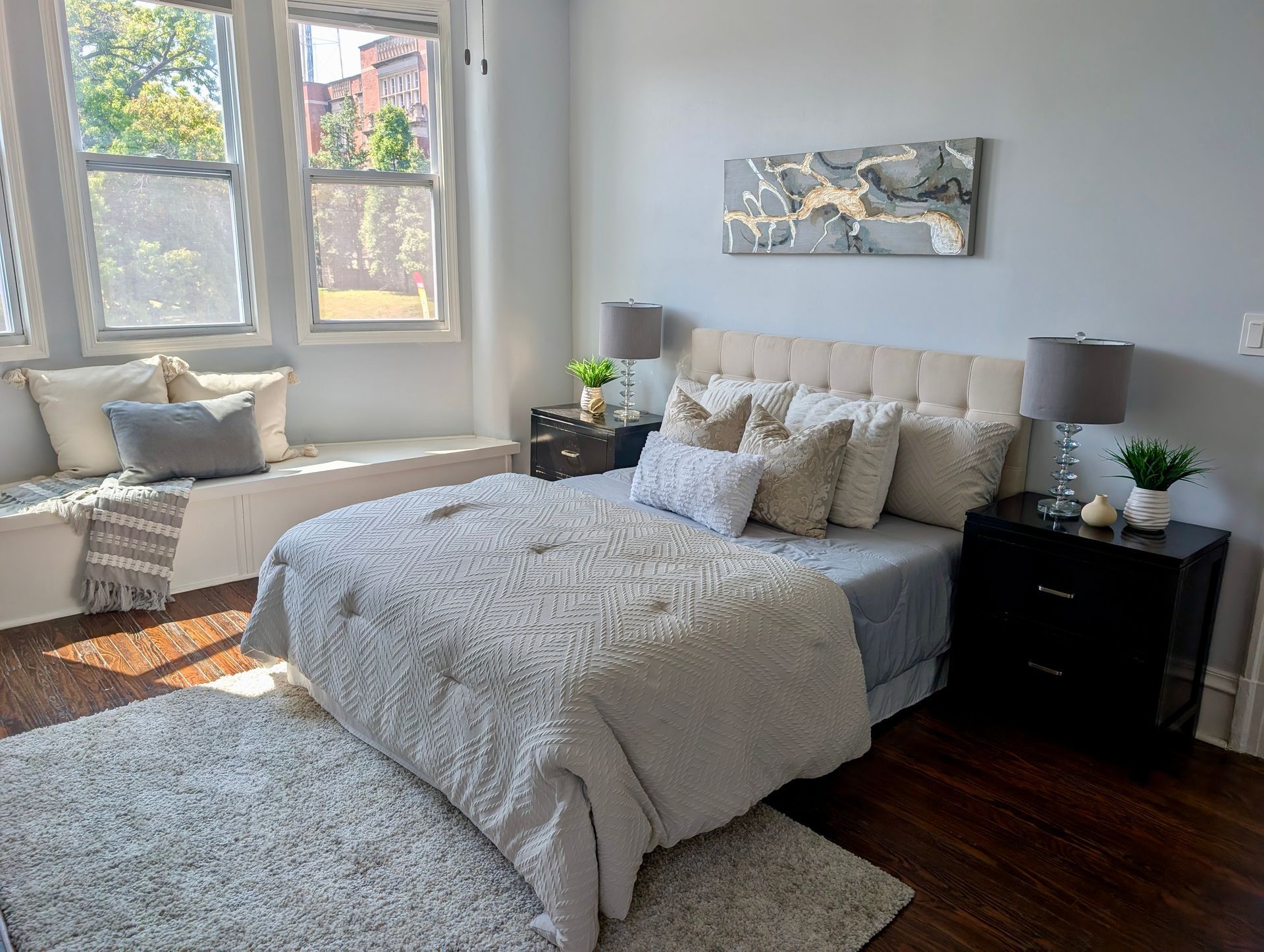 Bedroom with a window seat, bed, and two nightstands. Light blue walls, gray and white bedding, wooden floor.