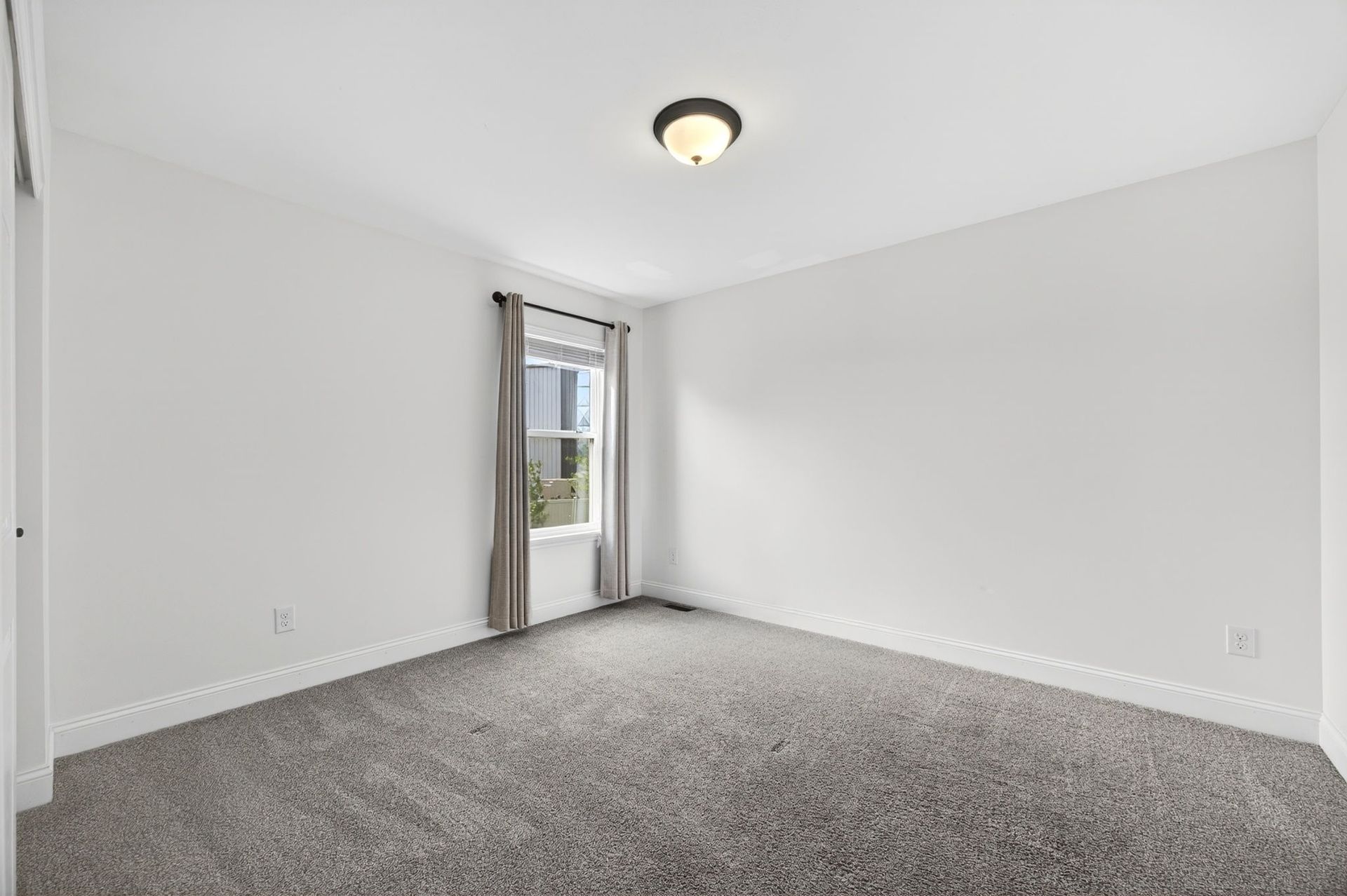 A bright, empty bedroom with white walls, gray wall-to-wall carpeting, and a window with gray curtains.