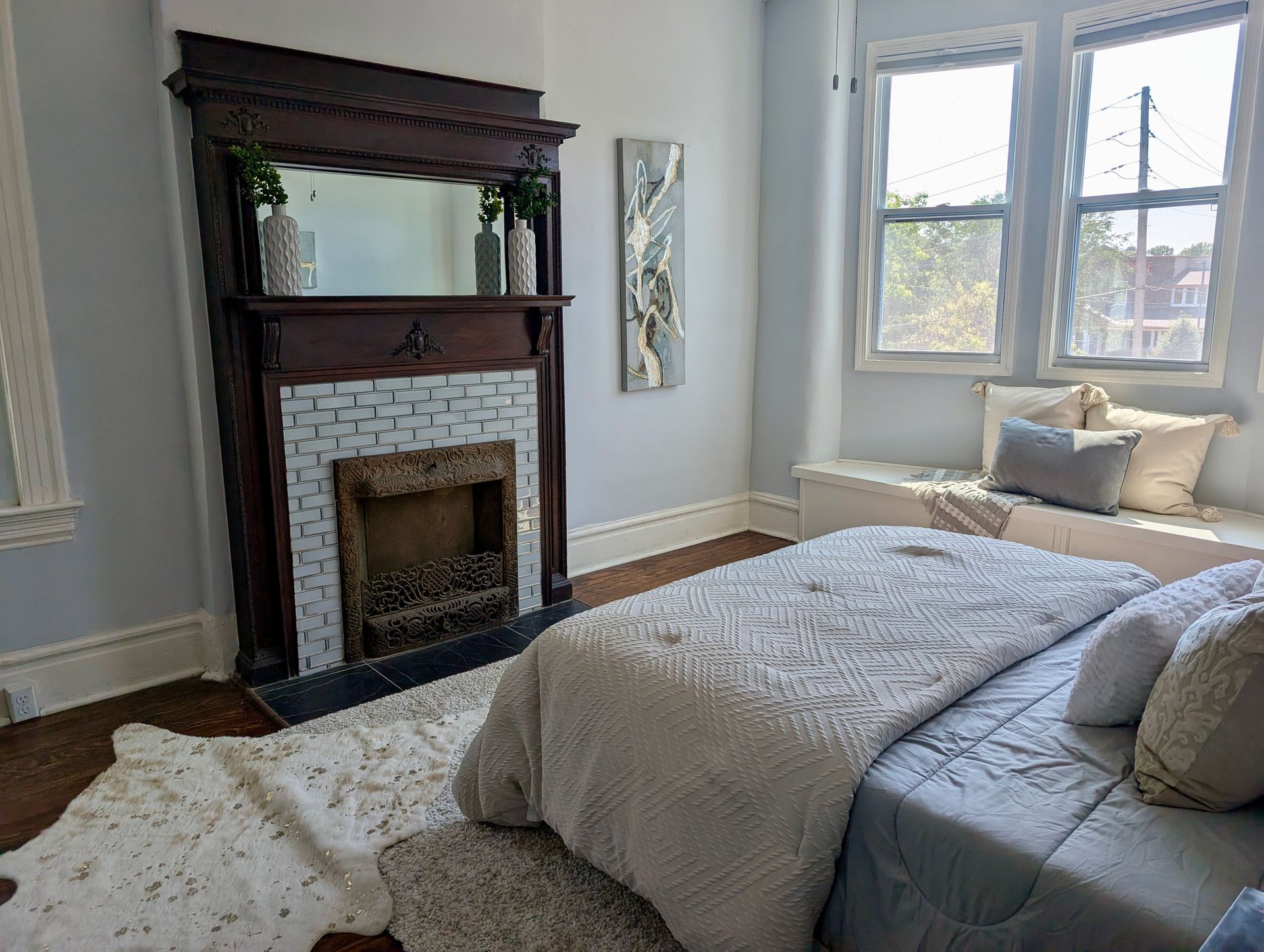 Bedroom with a bed, fireplace, and window seat. Light blue walls, white rug, and dark wooden fireplace surround.