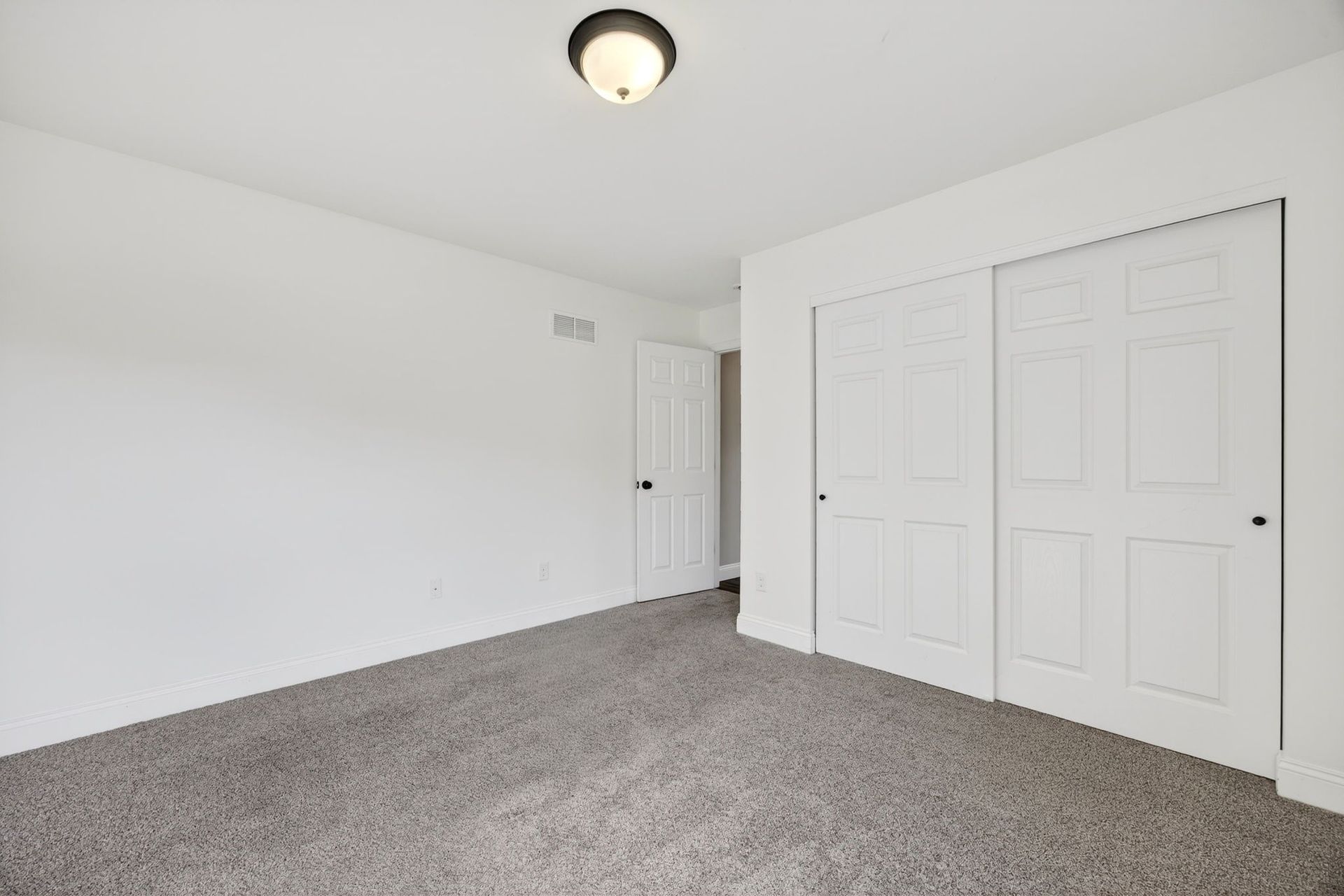 An empty bedroom with white walls, gray carpet, a white door, and a white sliding closet door under a ceiling light.