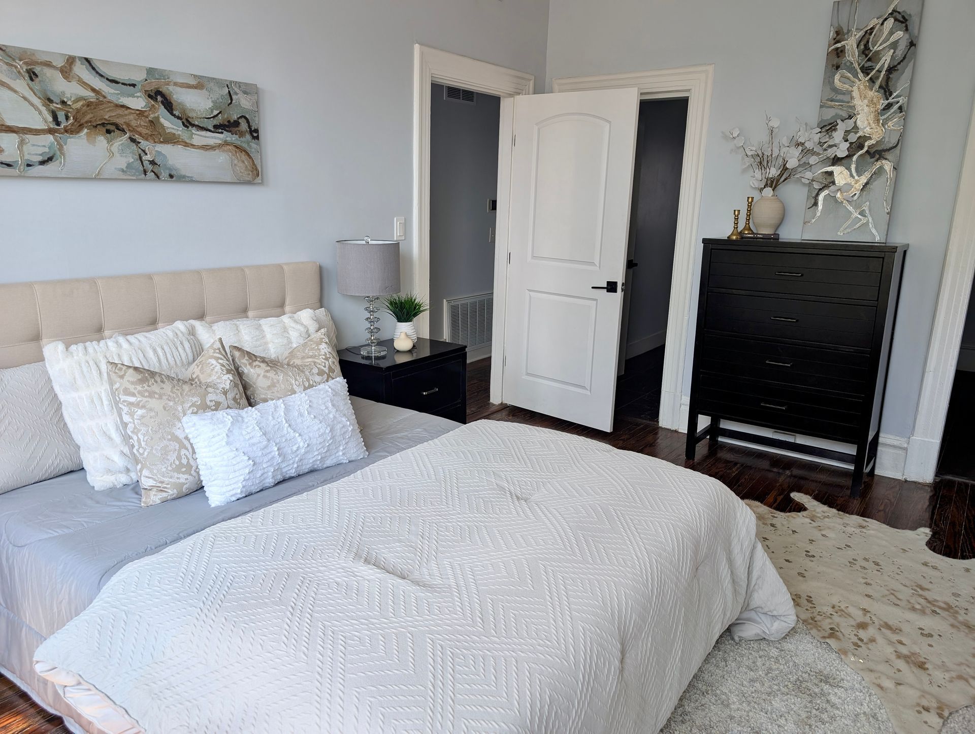 Bedroom with bed, dresser, artwork, and open door. Neutral colors with a rug on the floor.