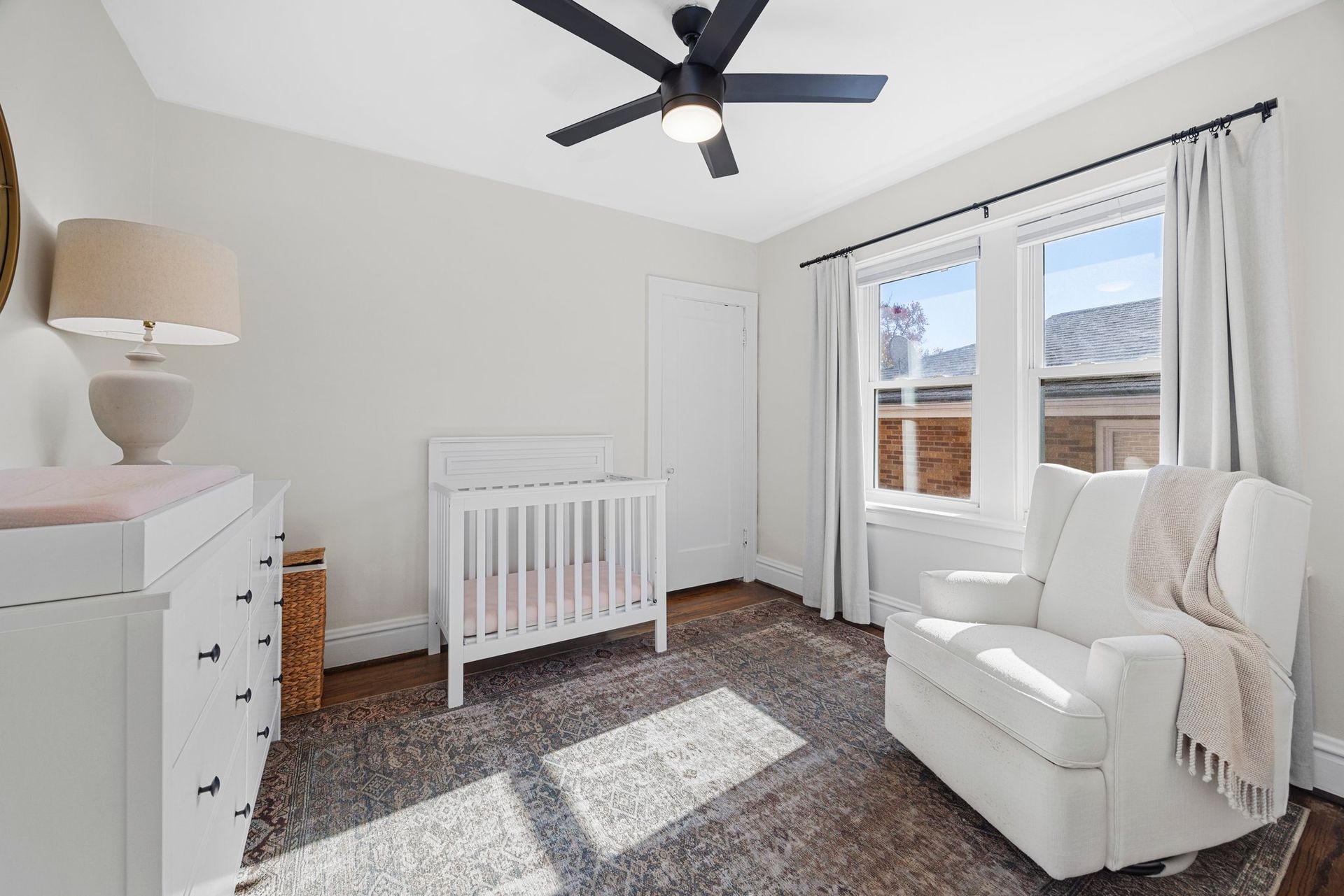 A nursery with a crib , chair , dresser and ceiling fan.