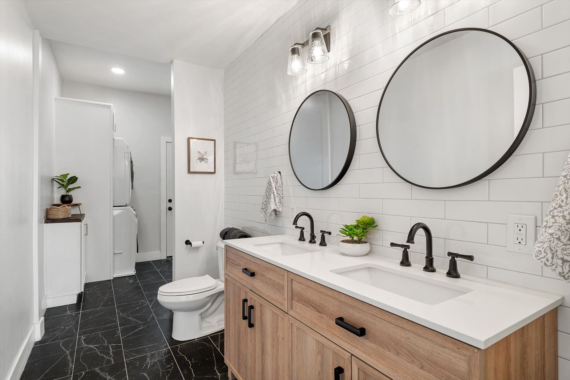 Modern bathroom with wood vanity, black hardware, round mirrors, white tile, and black marble floor.