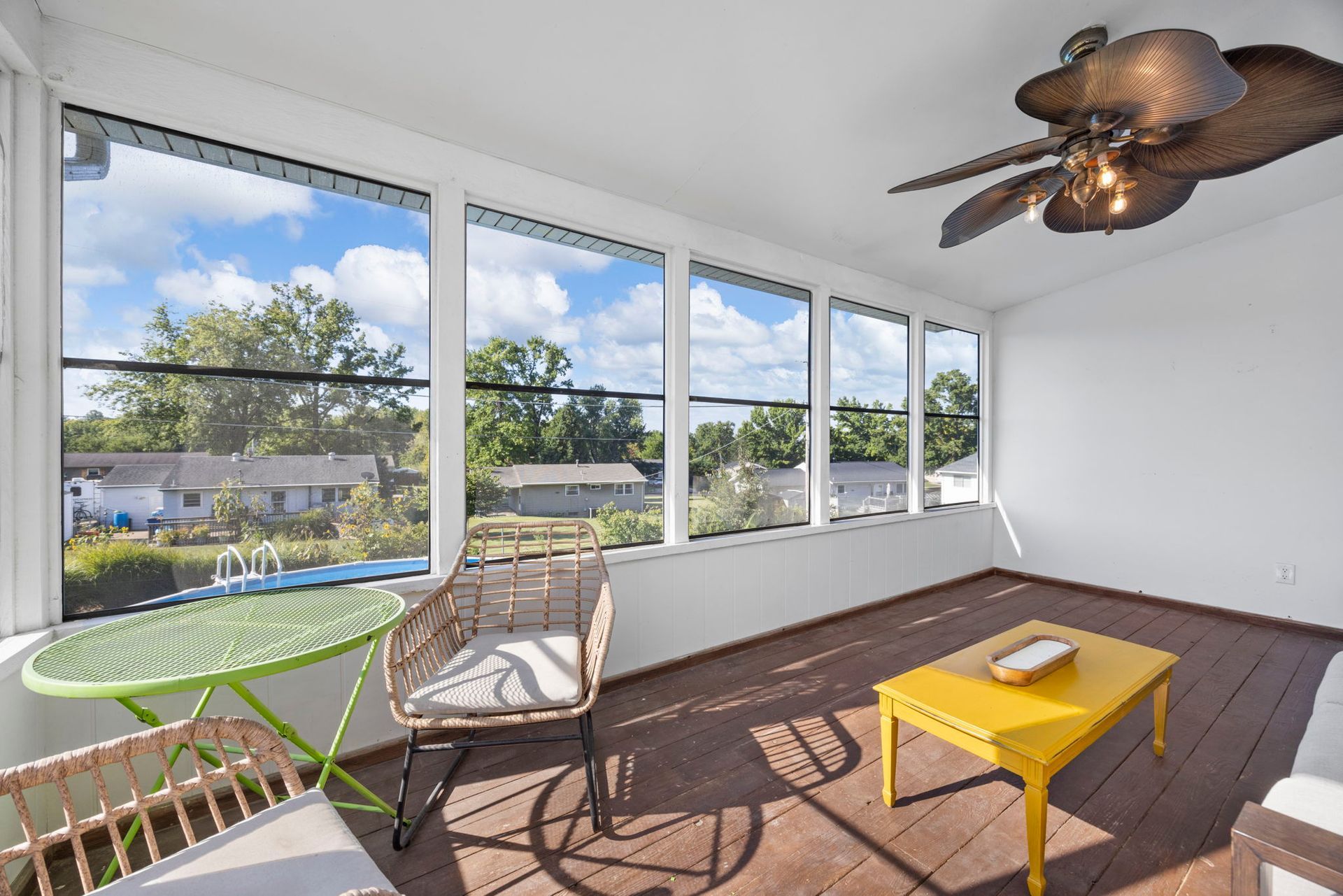 Sunroom with large windows, green table, woven chair, yellow coffee table, ceiling fan, and view of homes and sky.