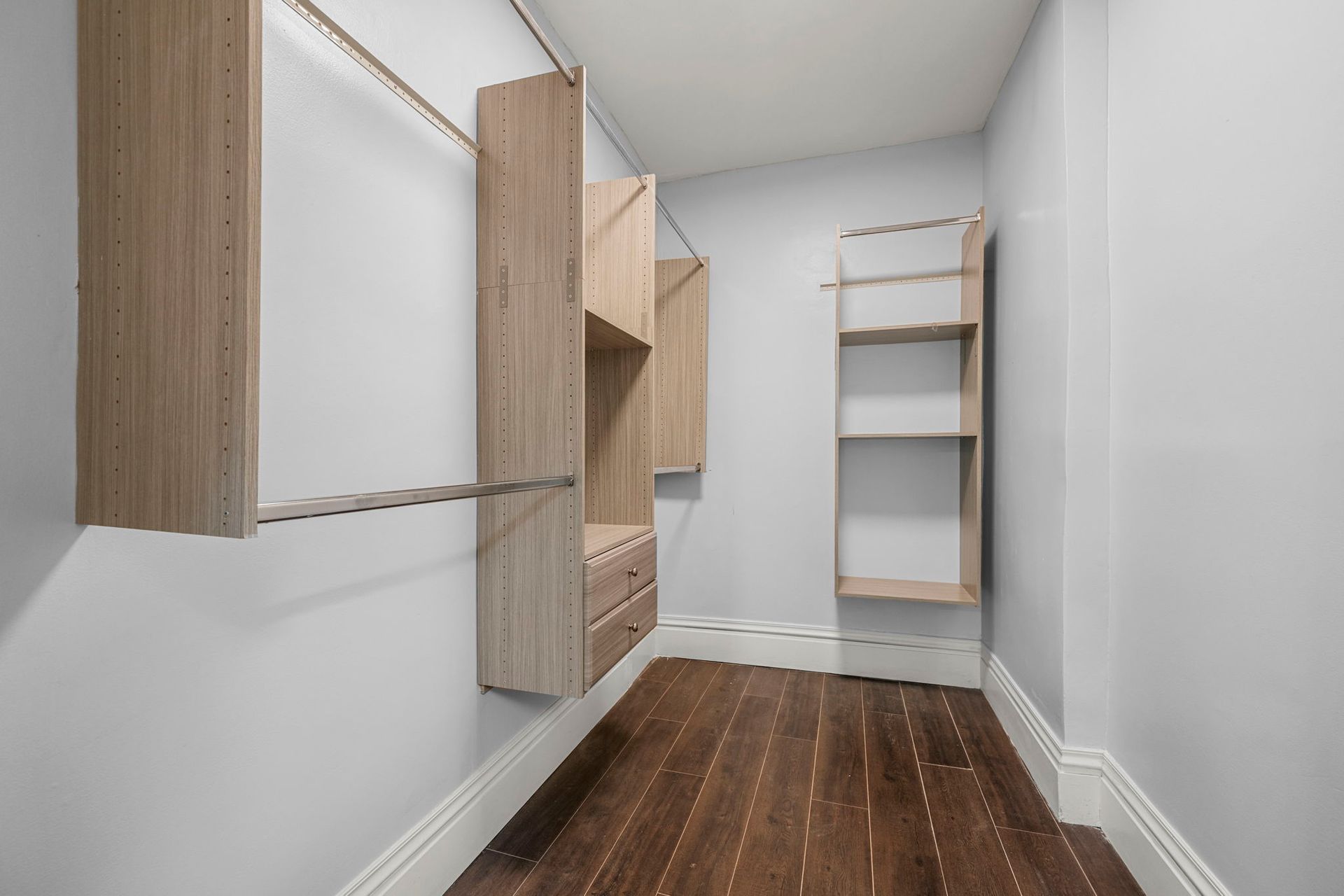 Empty walk-in closet with light wood shelves and hanging rods against gray walls. Dark wood floor.