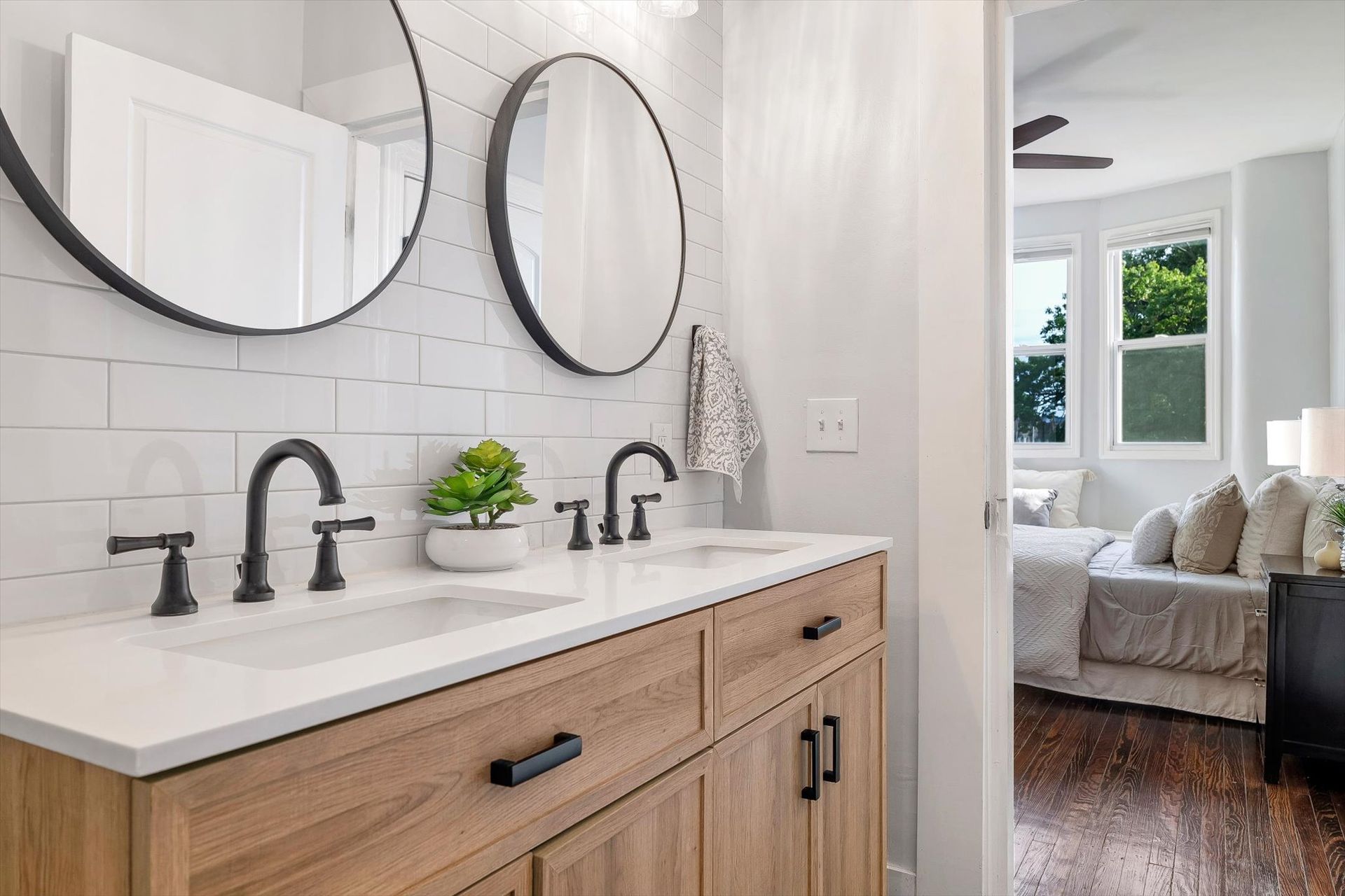 Bathroom with double sinks, wood vanity, subway tile, and two round mirrors; bedroom visible in background.