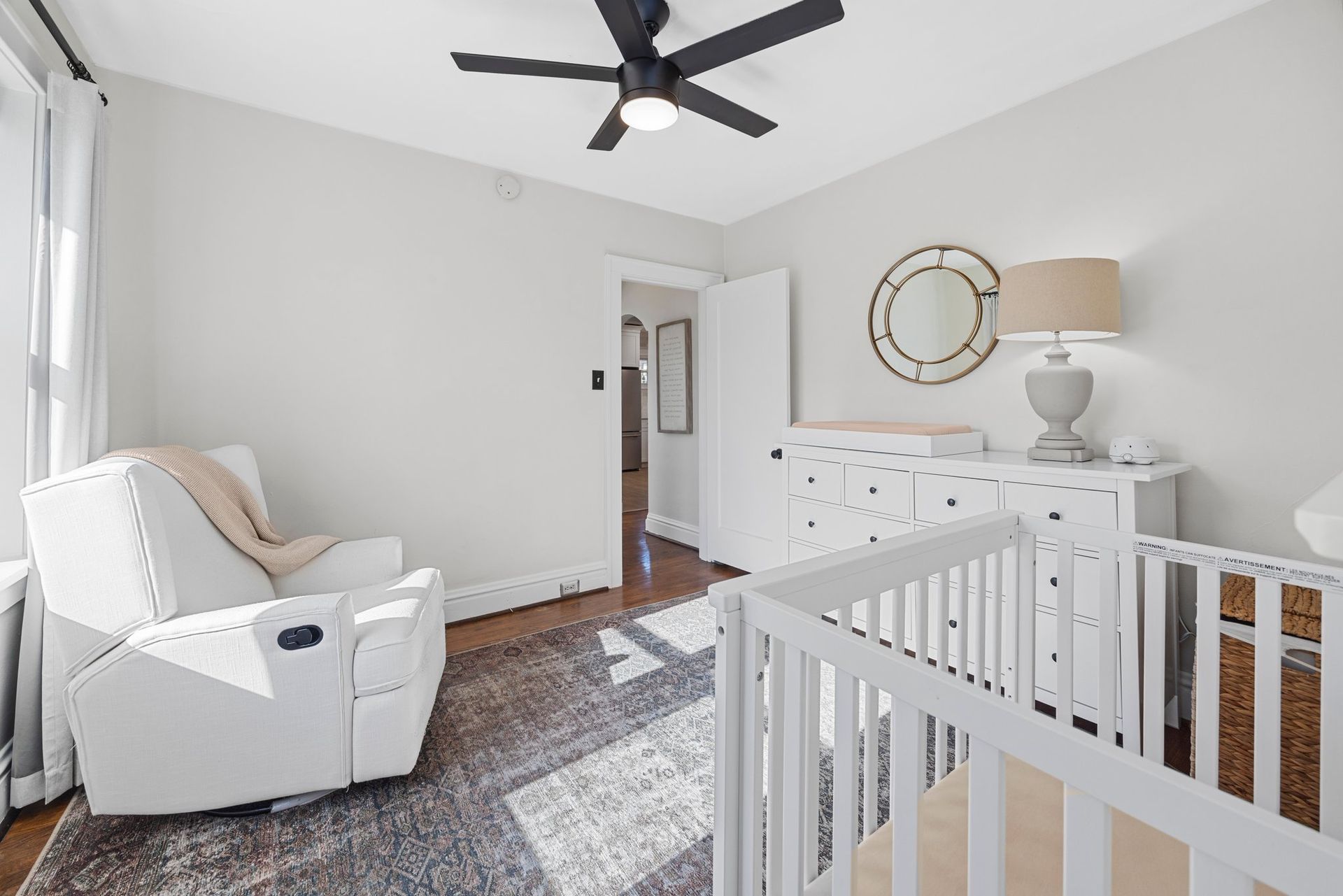 A nursery with a crib , chair , dresser and ceiling fan.
