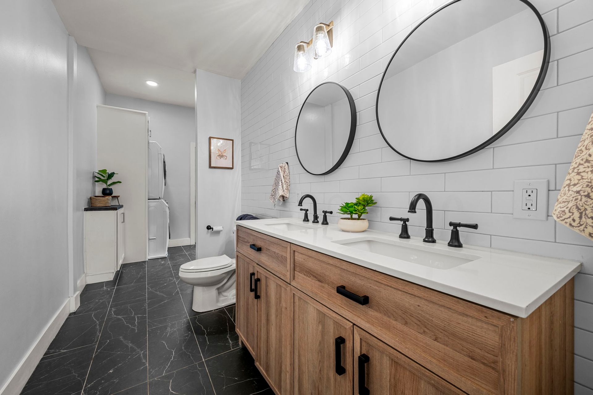 Bathroom with wooden vanity, white countertop, black fixtures, two round mirrors, and black tiled floor.