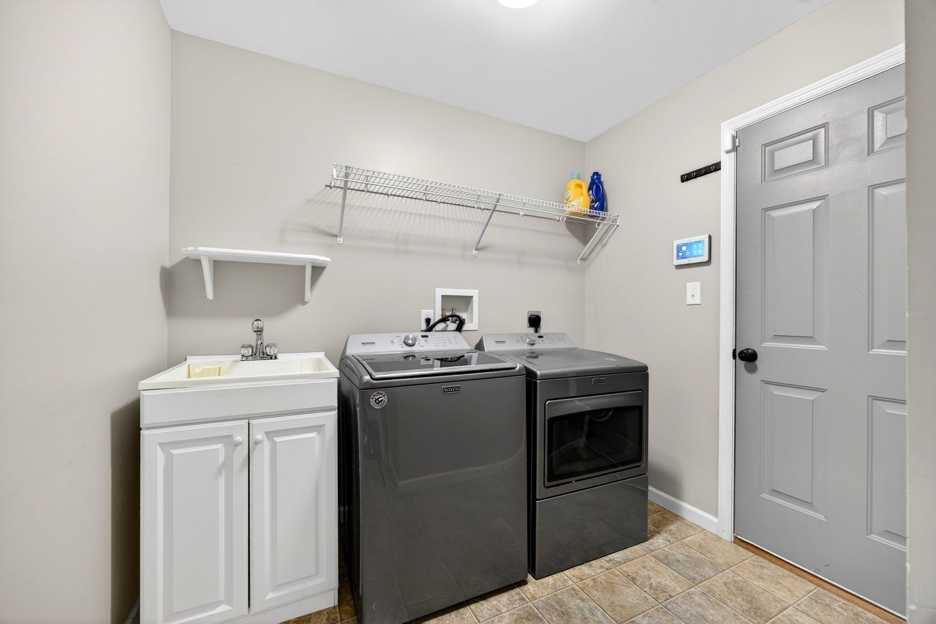 A laundry room featuring a white utility sink, grey washer and dryer, a wall-mounted shelf, and a grey door.