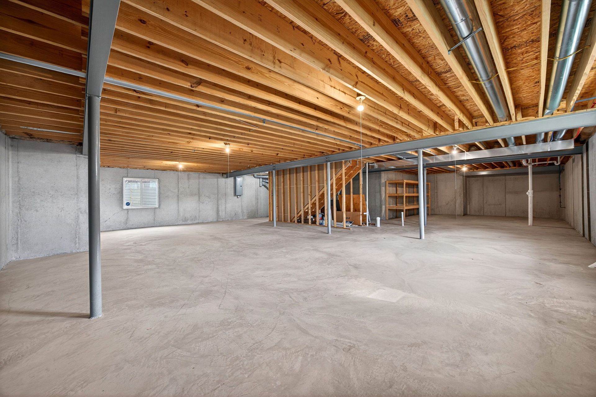 An unfinished basement with exposed wooden ceiling joists, concrete walls, a cement floor, and support poles.