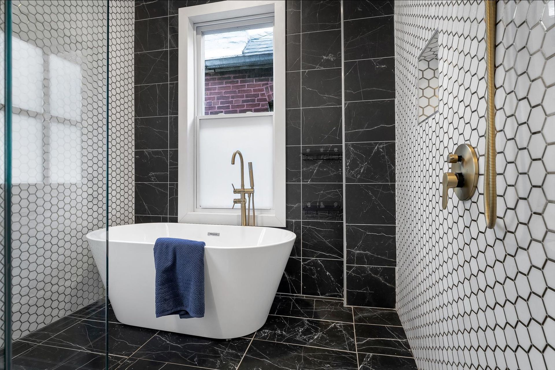 Modern bathroom with black marble tiles, white tub, and window with bronze fixtures.